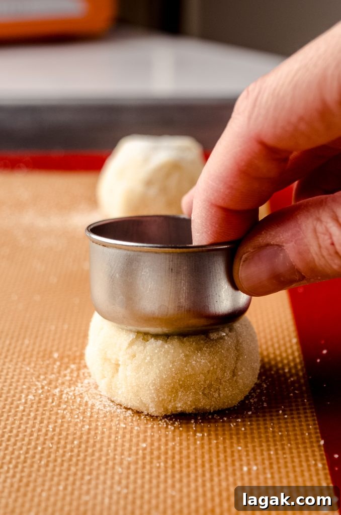 Cream Cheese Drop Cookies – Soft, Sugar Coated Treats 10 Someone is using a measuring cup to flatten a cookie dough ball on a baking sheet.