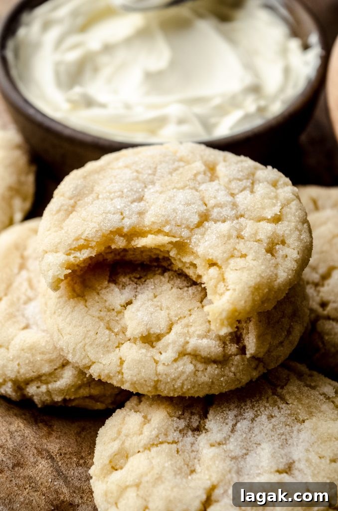 Cream Cheese Drop Cookies – Soft, Sugar Coated Treats 13 Cream cheese sugar cookies on a surface and a bite taken out of the one on the top with a bowl of cream cheese in the background.