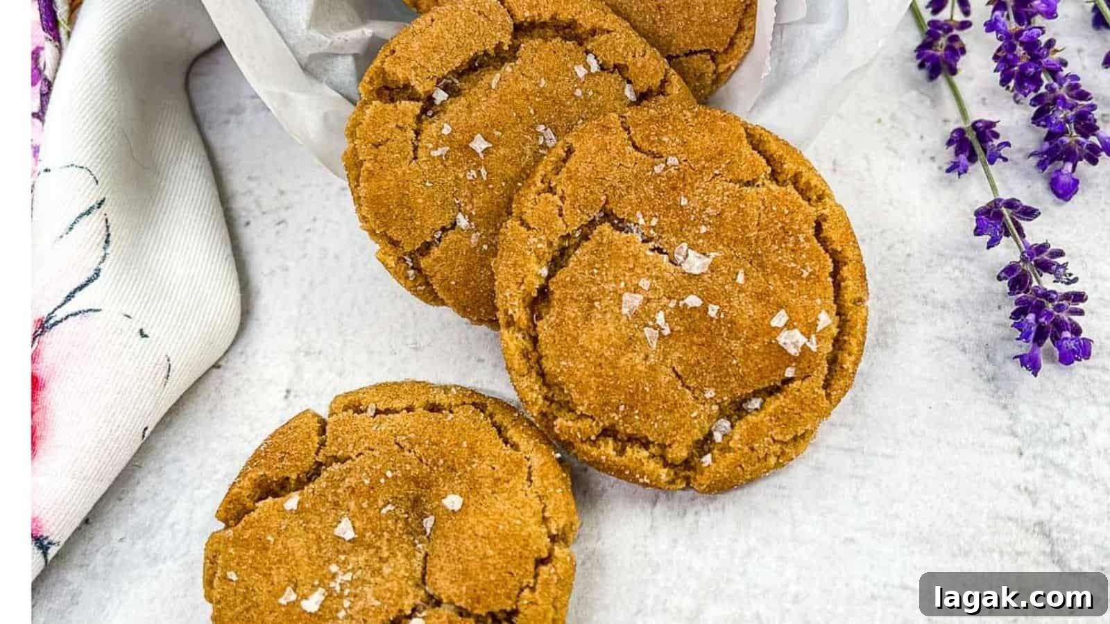 27 Cozy Fall Desserts That Taste Like Brown Sugar, Butter, and Memories 5 Three brown butter snickerdoodle cookies arranged on a white surface, sprinkled with sea salt flakes, with a white cloth and purple flowers on the side.