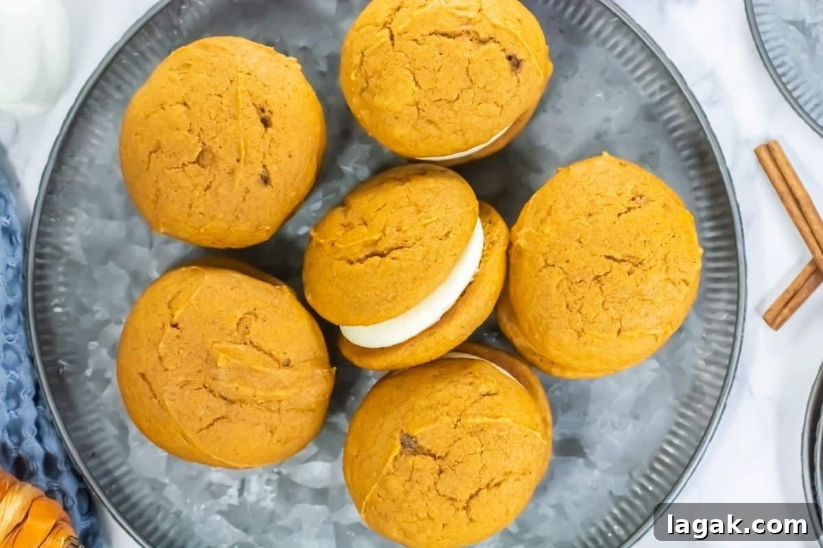27 Cozy Fall Desserts That Taste Like Brown Sugar, Butter, and Memories 10 Six pumpkin whoopie pies with cream filling are arranged on a gray plate, viewed from above. Two cinnamon sticks are visible at the edge of the plate.
