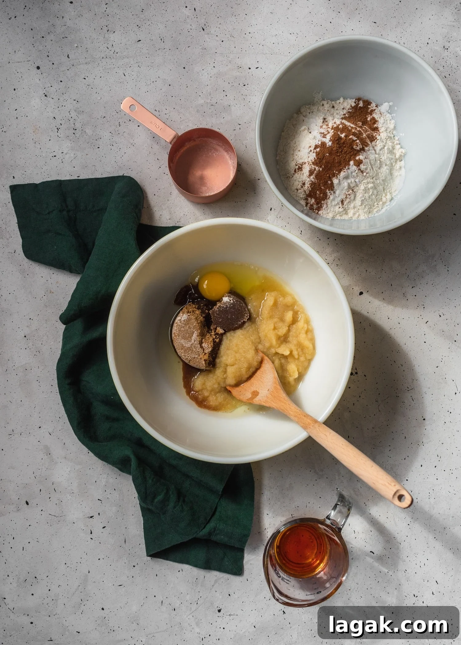 A white mixing bowl with eggs, sugar, applesauce and oil with a wooden spoon sticking out, surrounded by a bowl of flour, a bronze mixing cup, and a green linen on a grey counter.