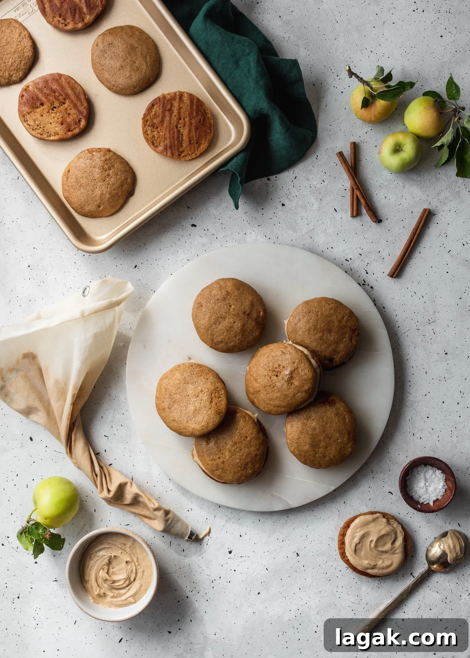 An overhead shot of a white plate filled with apple cider whoopie pies, surrounded by a baking pan, apples, cinnamon, and a piping bag.