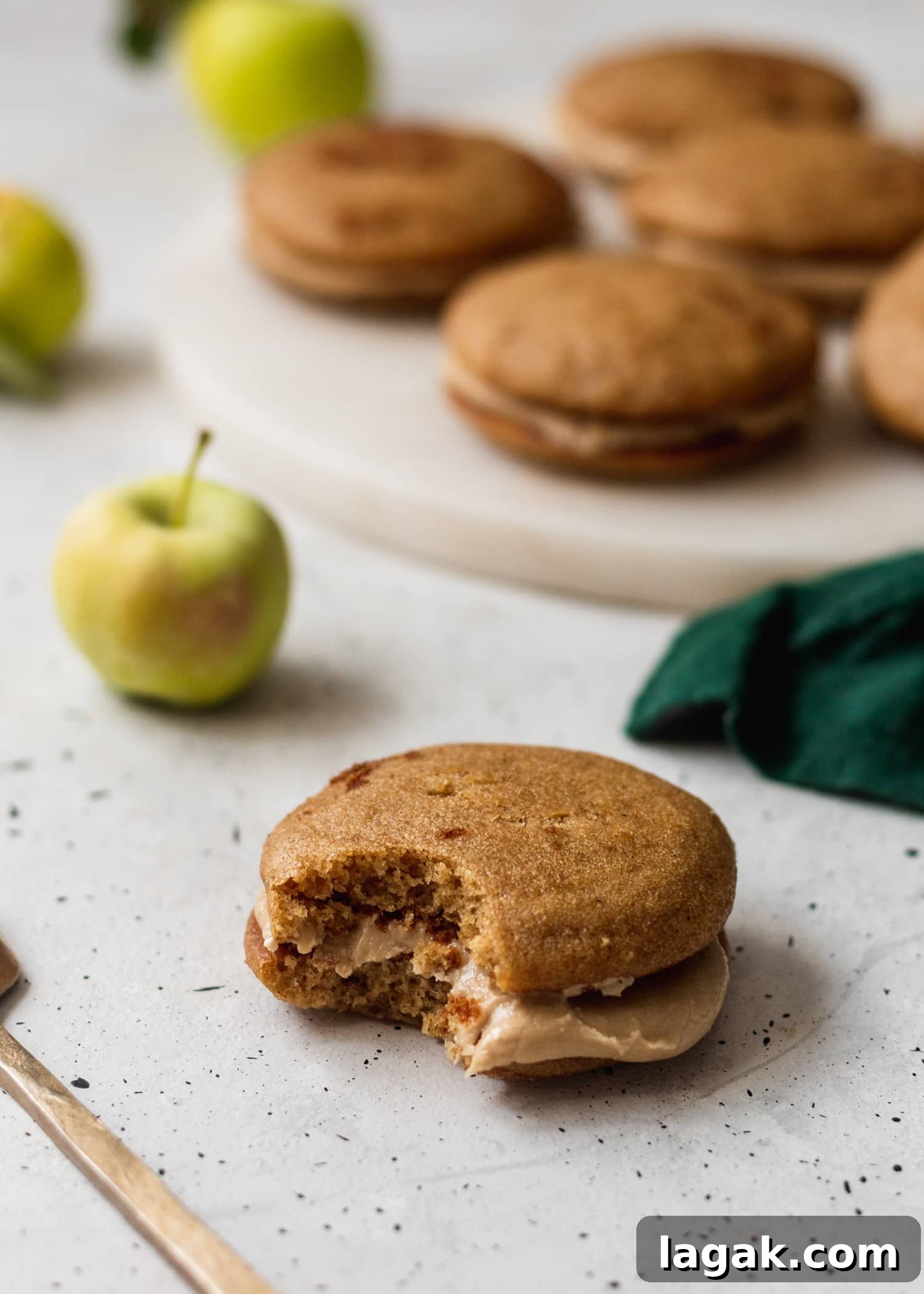 A close up shot of an apple cider whoopie pie with a bite taken out of it sitting on a white speckled table with a plate of apple cider whoopie pies, a green linen, and an apple in the background.