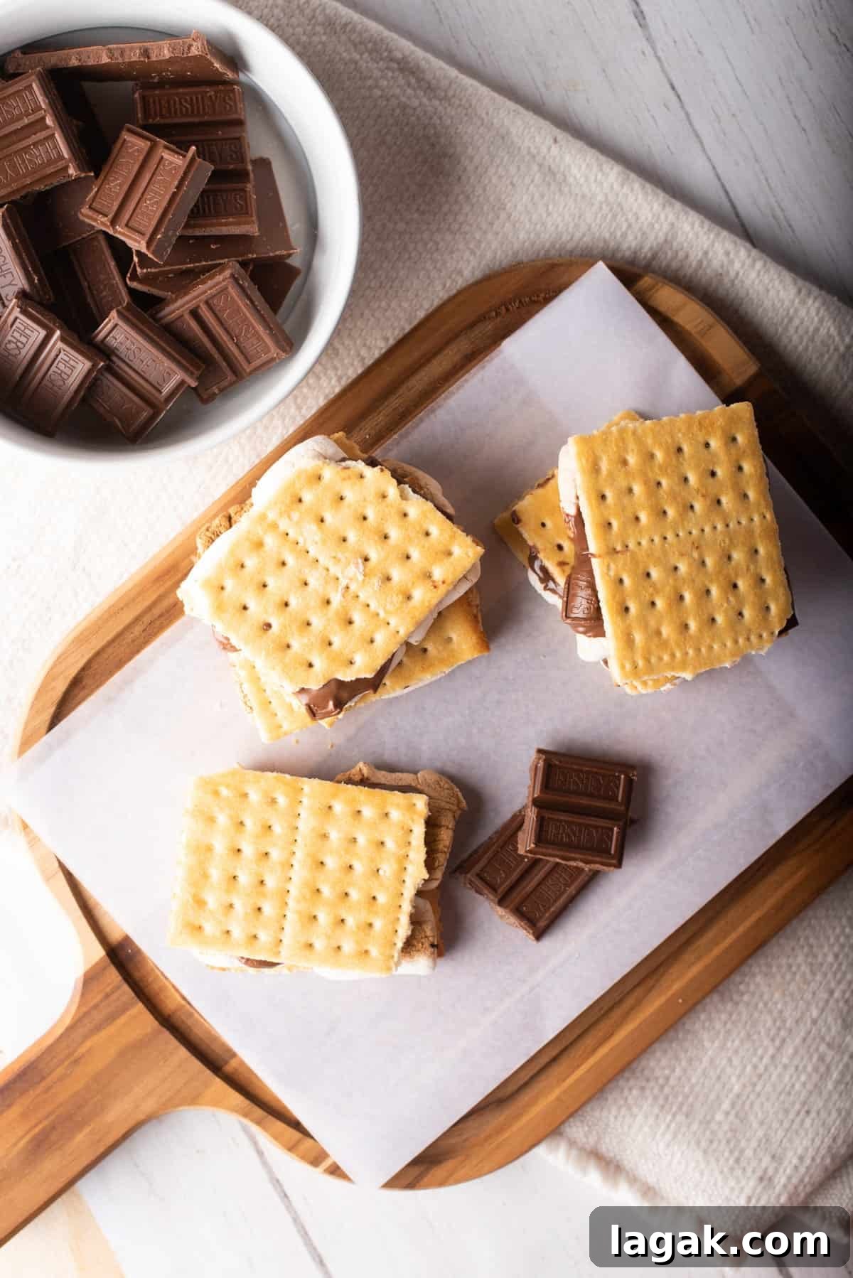 Air Fryer S'mores: Quick Indoor Campfire Dessert Recipe 9 Overhead view of three air-fried s'mores placed on a wooden board next to a bowl of chocolate pieces.