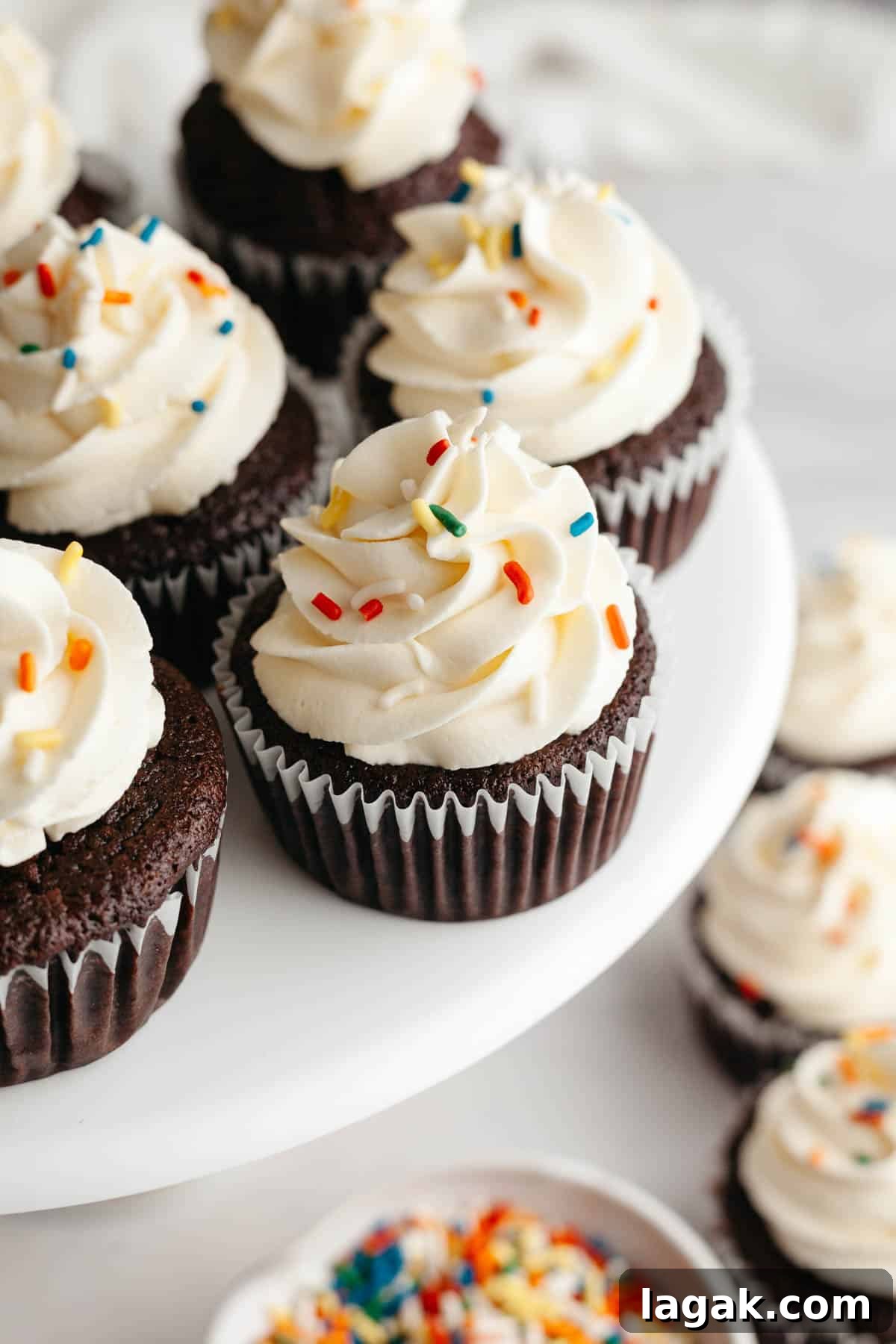 Moist Chocolate Cupcakes with Cream Cheese Frosting Recipe 2 Chocolate cupcakes with a white frosting and rainbow sprinkles on a cake stand.