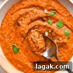 A wide, ceramic bowl of bright orange romesco sauce, garnished with chopped almonds and fresh parsley. A spoon is dipping into the center of the bowl to show the smooth yet rustic texture.