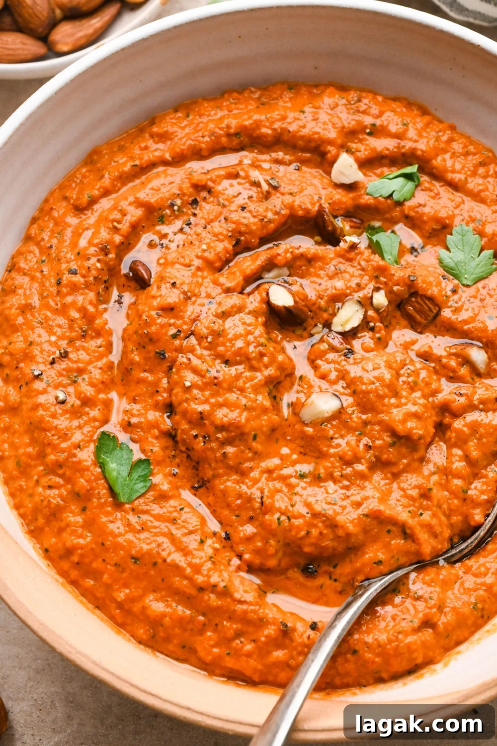 10-Minute Rustic Romesco Sauce (Dairy-Free) 3 A wide, ceramic bowl of bright orange romesco sauce, garnished with chopped almonds and fresh parsley. A spoon is angled into the sauce on the side of the bowl.