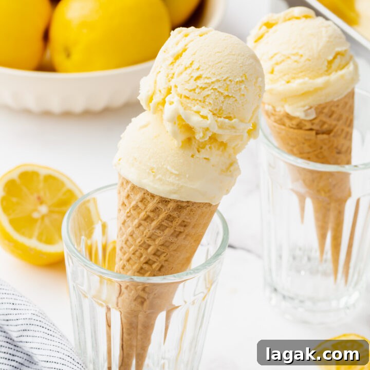 Two sugar cones topped with light yellow lemon ice cream, being held up in glass drinking glasses. A bowl of lemons is in the background.