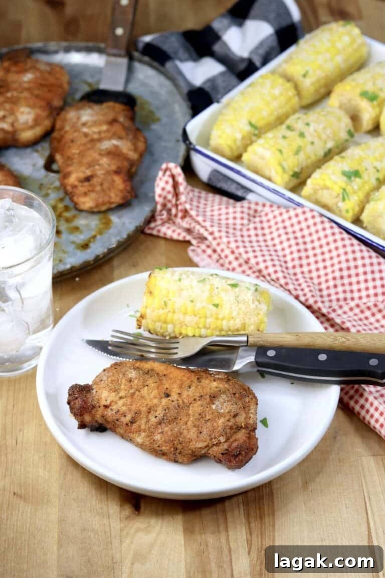 a grilled pork chop on a white plate next to grilled corn and silverware.