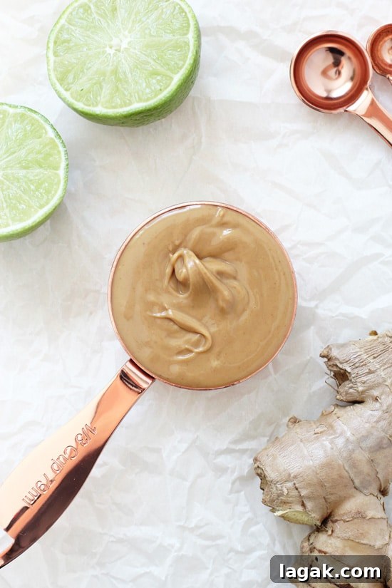 Filled with creamy peanut butter, fresh ginger, garlic, coconut milk and lime juice, this easy homemade peanut sauce is packed with Thai inspired flavor! Ready in 15 minutes or less! A measuring cup filled with peanut butter and fresh ginger and limes to the side.