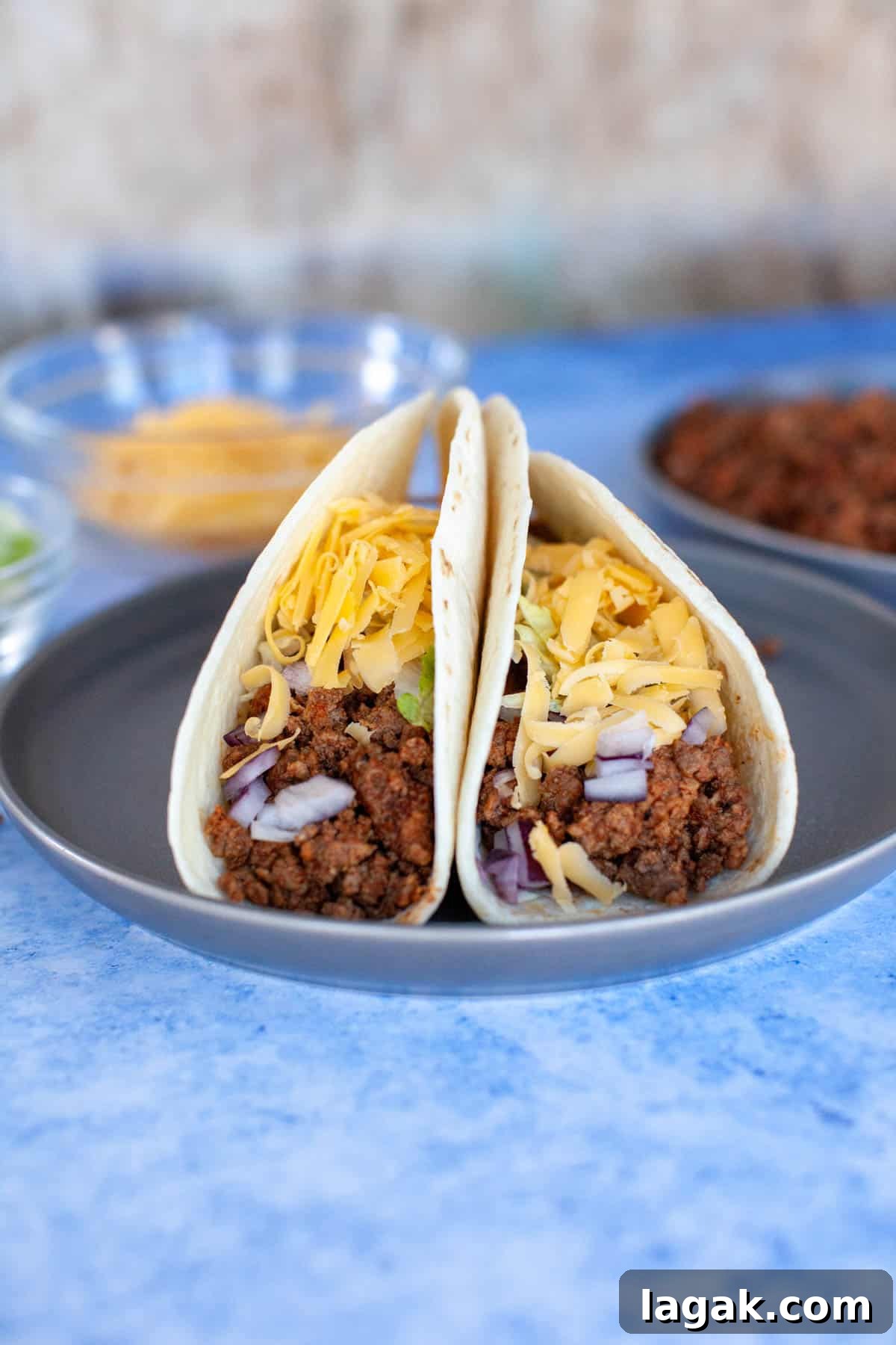Authentic Ground Beef Tacos Recipe for Weeknight Dinners 9 Two ground beef tacos on a plate with cheese, chopped red onion and shredded lettuce.