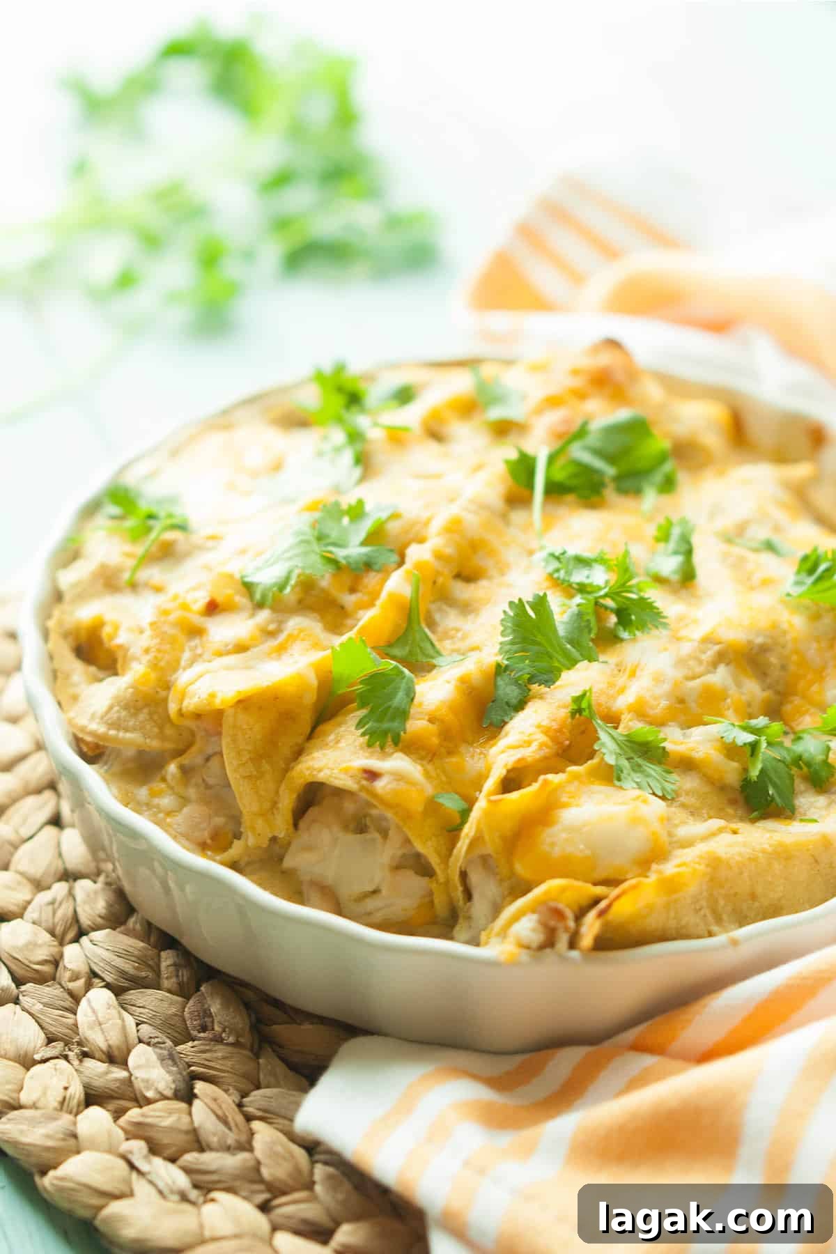 Gluten-Free Cream Cheese Chicken Enchiladas (Video Recipe) 2 Cream cheese gluten-free chicken enchiladas in a white pan with cilantro on top on a rattan mat with a white and orange linen napkin.