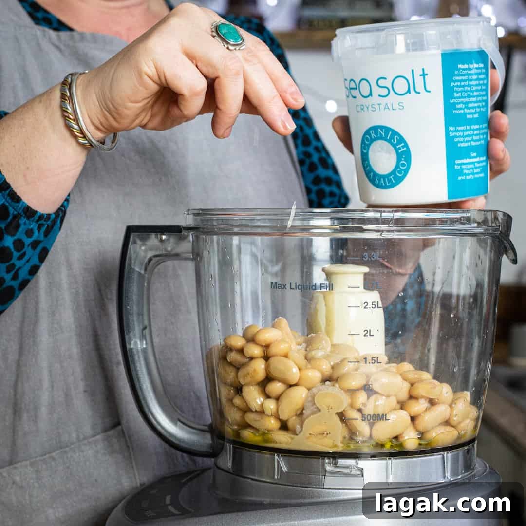 Creamy Vegan Butter Bean Dip — Protein-Packed Snack Recipe 7 Woman sprinkling salt into a mixer of hummus ingredients