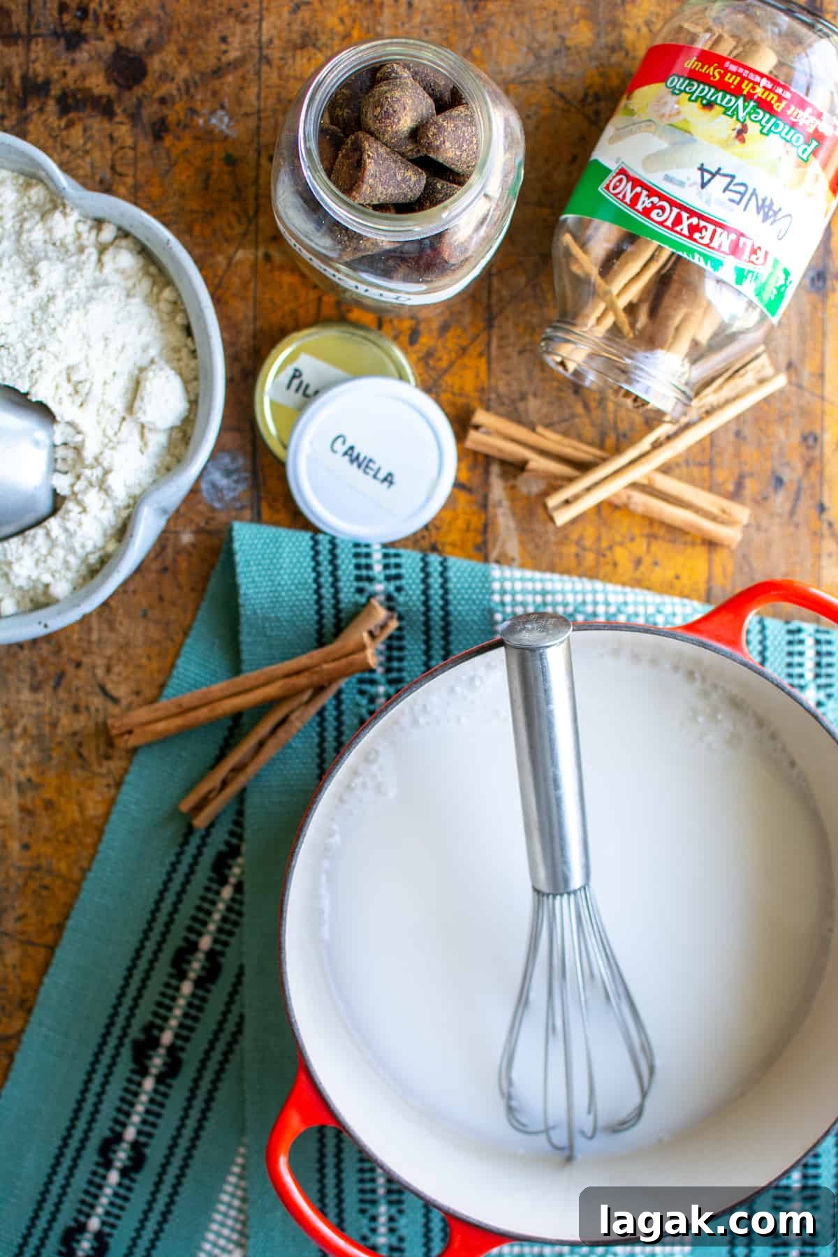 Vegan Atole: Warm Cinnamon Corn Drink Recipe 4 Ingredients to make atole on a wood table, including, masa harina, piloncillo, canela, and a large red pot with milk and a whisk in it.
