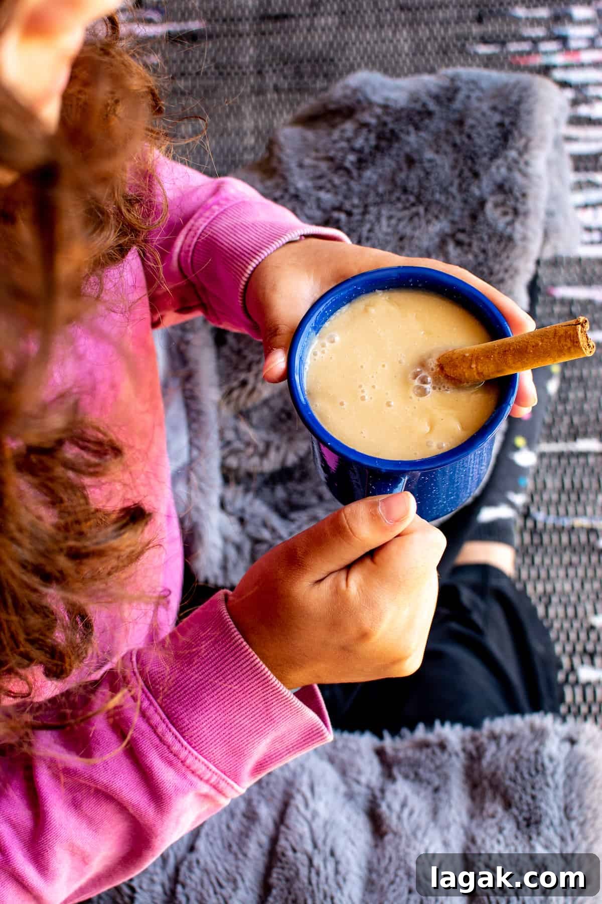 Vegan Atole: Warm Cinnamon Corn Drink Recipe 6 A girl in a pink sweater with a blanket wrapped around her holding a blue mug filled with atole and a cinnamon stick sitting in the mug.