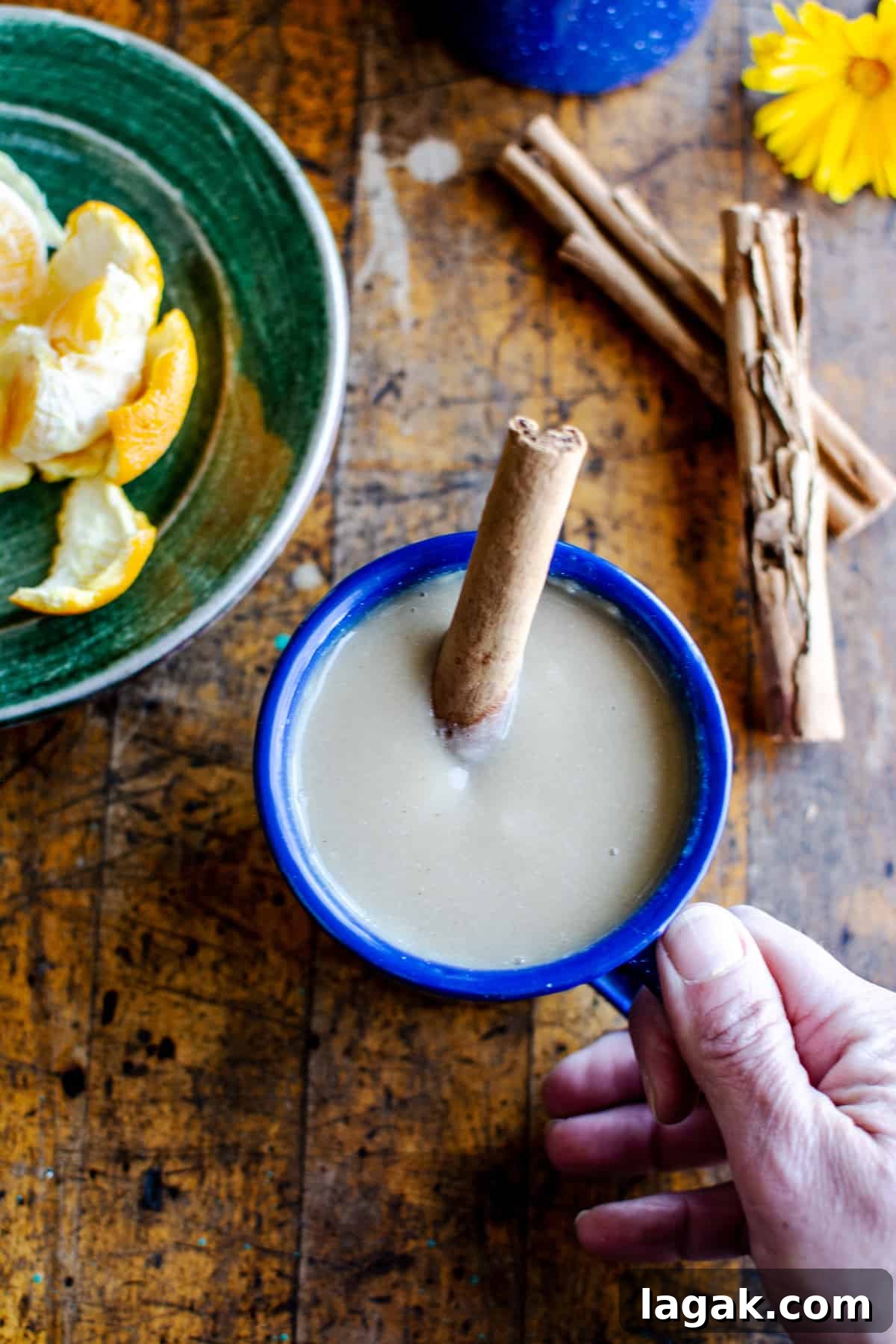 Vegan Atole: Warm Cinnamon Corn Drink Recipe 7 A blue mug of atole with a cinnamon stick in it and a hand holding the handle of the mug.