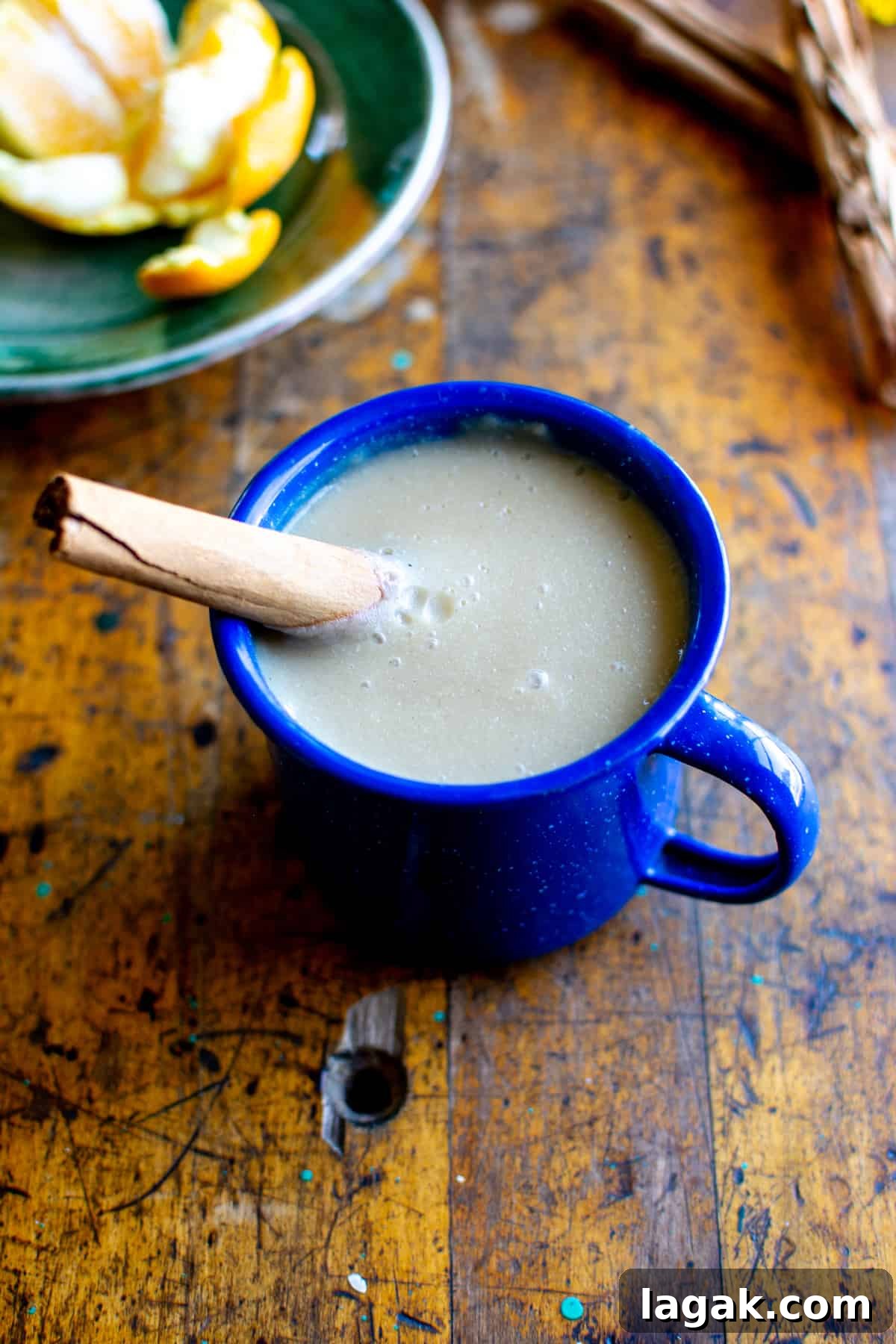 Vegan Atole: Warm Cinnamon Corn Drink Recipe 8 A mug of atole with a cinnamon stick sticking in the mug sitting on a wood table with a small plate of oranges behind it.