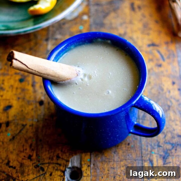 A mug of atole with a cinnamon stick sticking in the mug sitting on a wood table with a small plate of oranges behind it.