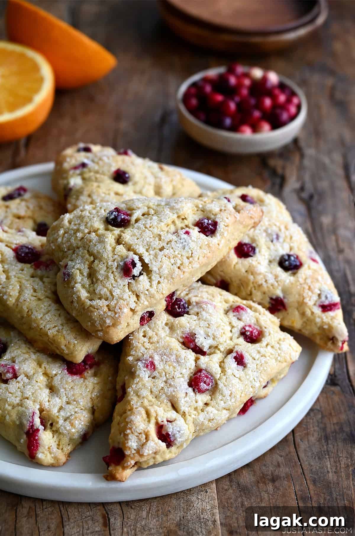 Cranberry Orange Scones with Sweet Citrus Glaze 5 Cranberry orange scones with coarse sugar on a serving plate near a bowl of fresh cranberries and an orange that's been cut in half.
