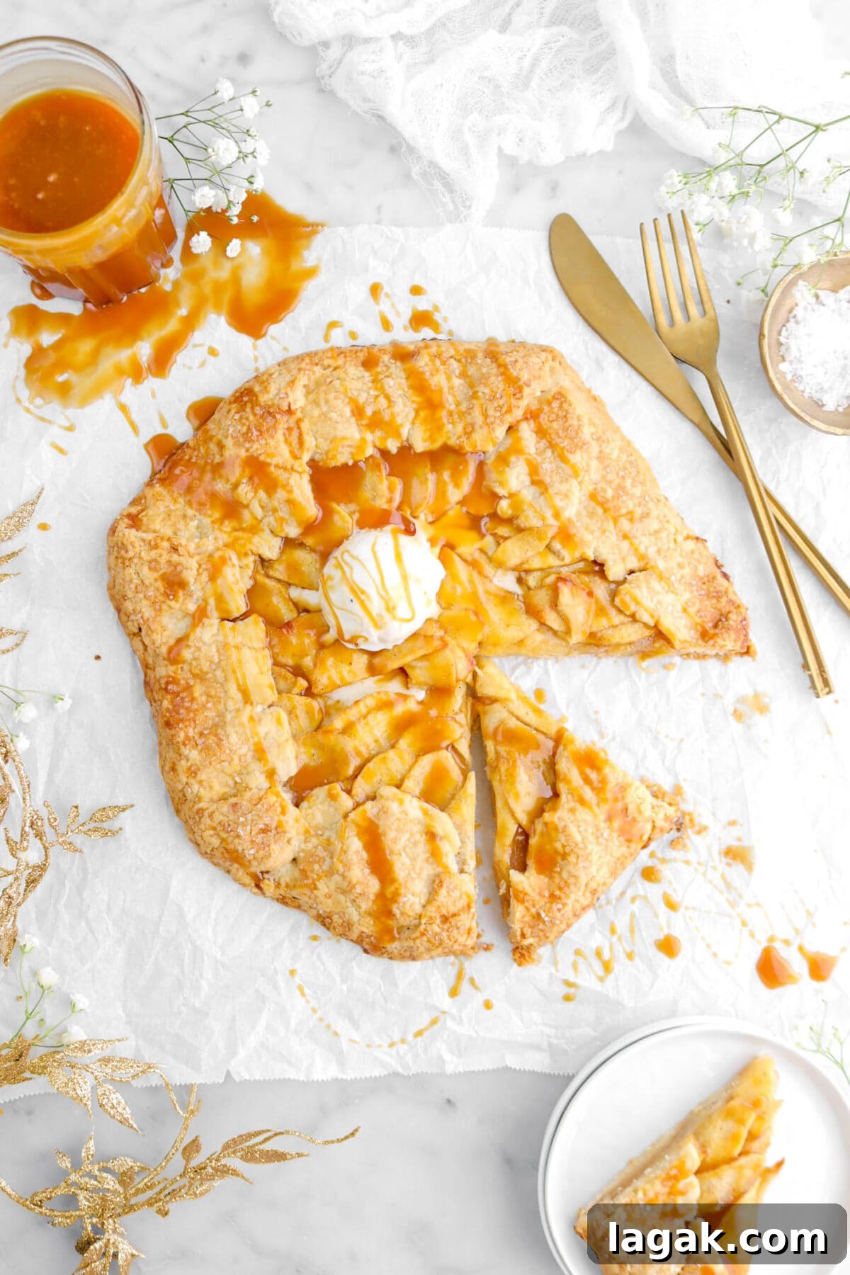 Caramel Apple Galette Recipe: Rustic Fall Dessert with Salty Caramel 2 caramel apple galette with scoop of ice cream on top on parchment paper with slice on plate beside, gold leaf garland and jar of caramel to the left side, and fork and knife to the right side