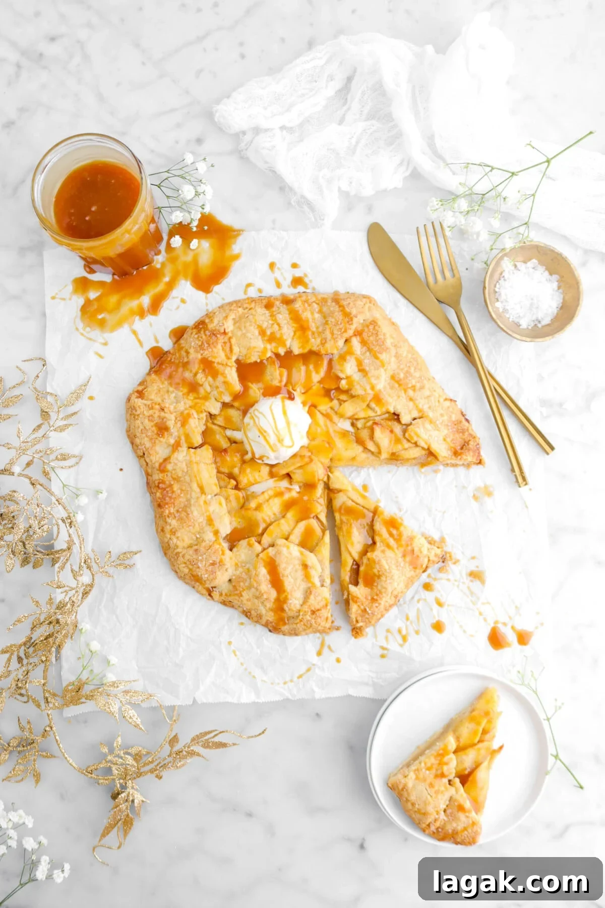 Caramel Apple Galette Recipe: Rustic Fall Dessert with Salty Caramel 5 overhead shot of caramel apple galette with slice beside and second slice on white plate with gold garland beside, white flowers around, jar of caramel, and white cheese cloth on marble surface