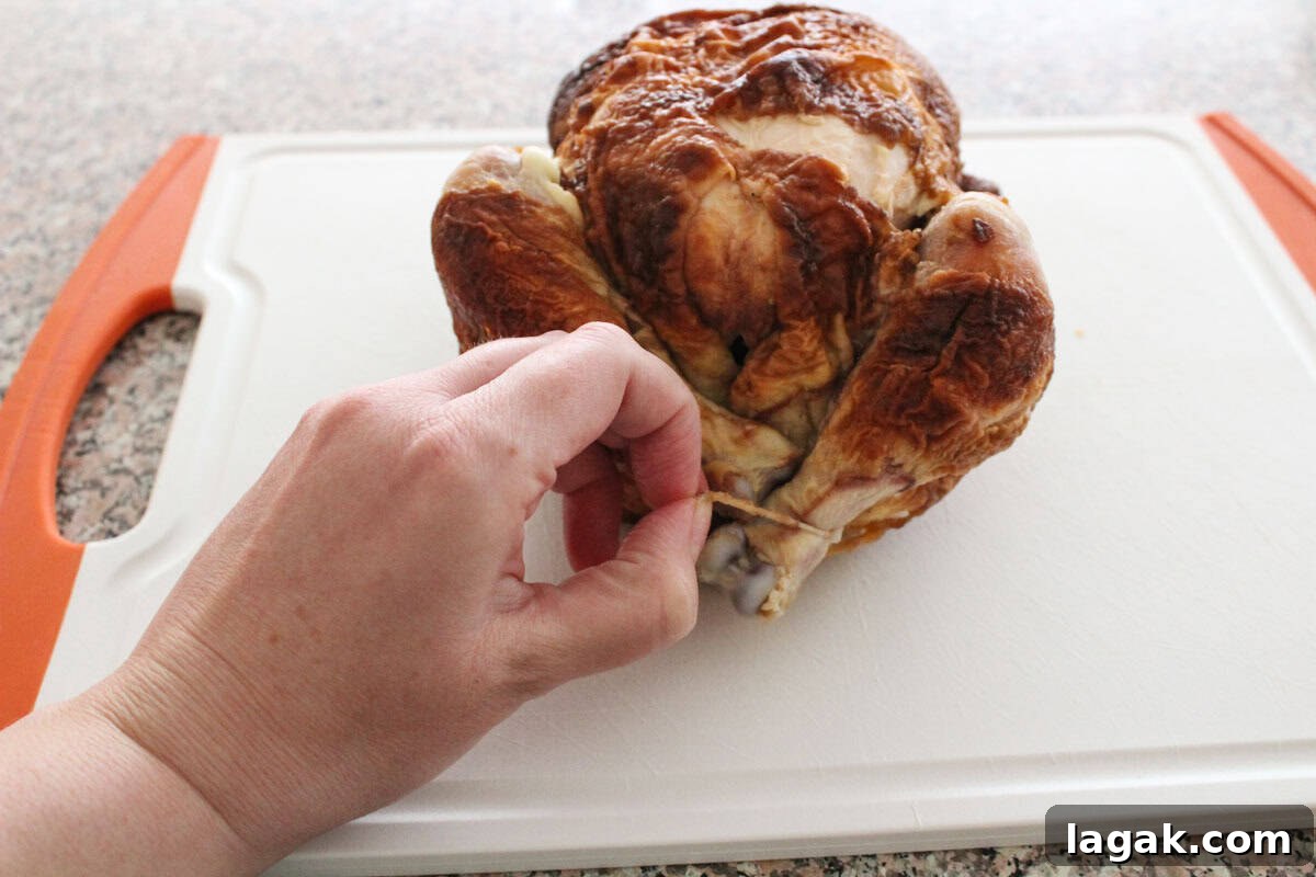How to Cut and Shred Rotisserie Chicken: Portioning & Freezing Tips 4 A hand unties kitchen twine from the legs of a cooked rotisserie chicken resting on a white cutting board with orange handles.