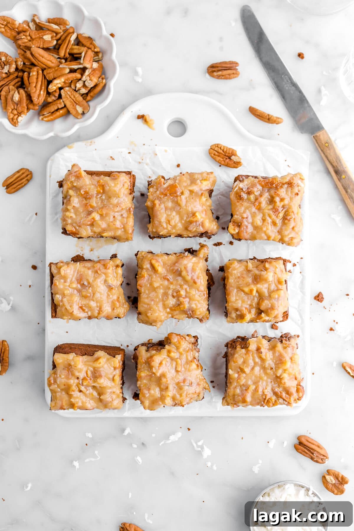 overhead shot of german chocolate brownies on white tray with a knife, bowl of pecans, and measuring cup of shredded coconut around.