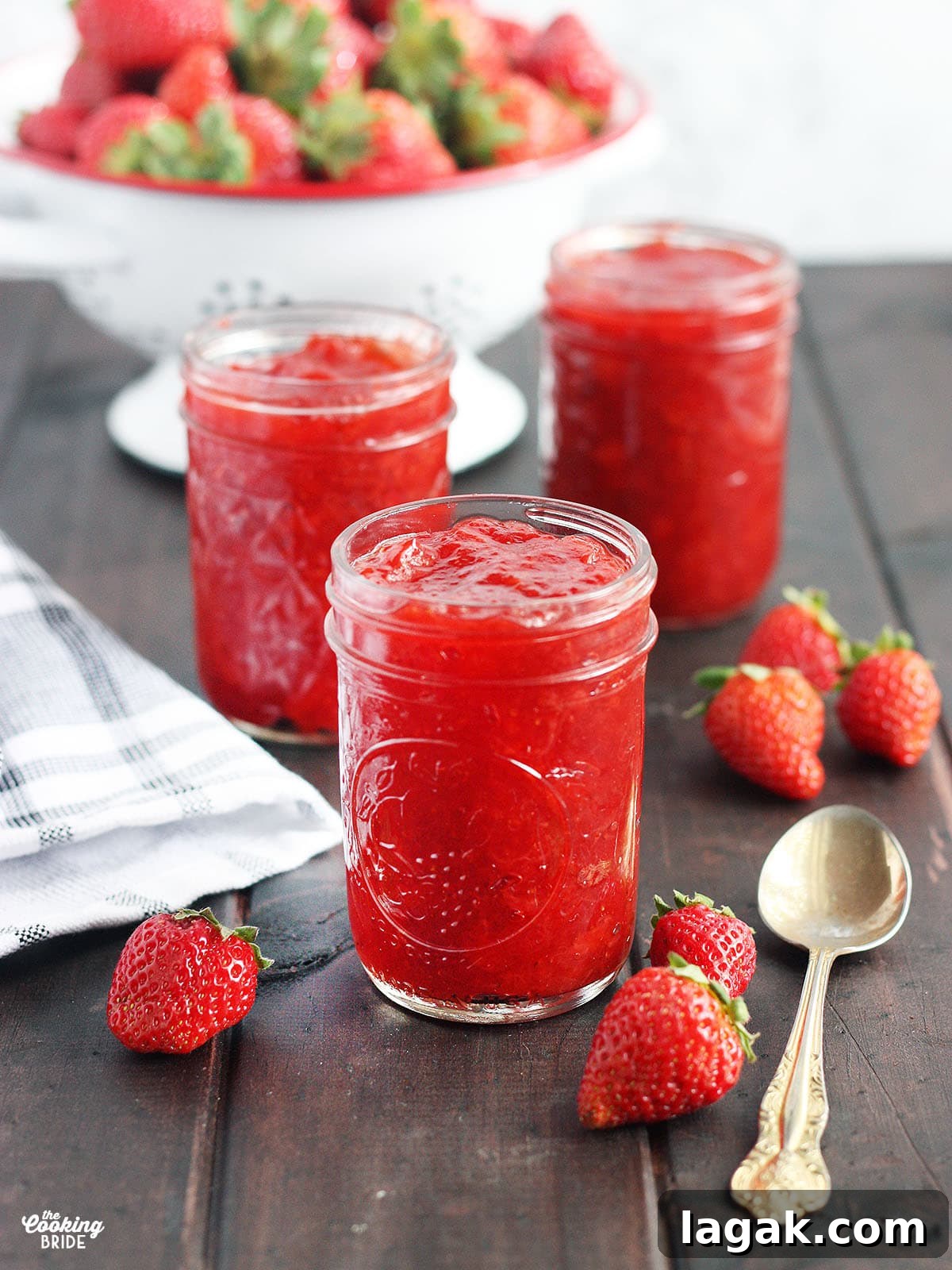 Strawberry Jam Recipe with Pectin: Smooth, Spreadable Preserves 2 three jars of homemade strawberry preserves on a wooden table with fresh strawberries and a spoon to the side, colander of fresh strawberries in the background