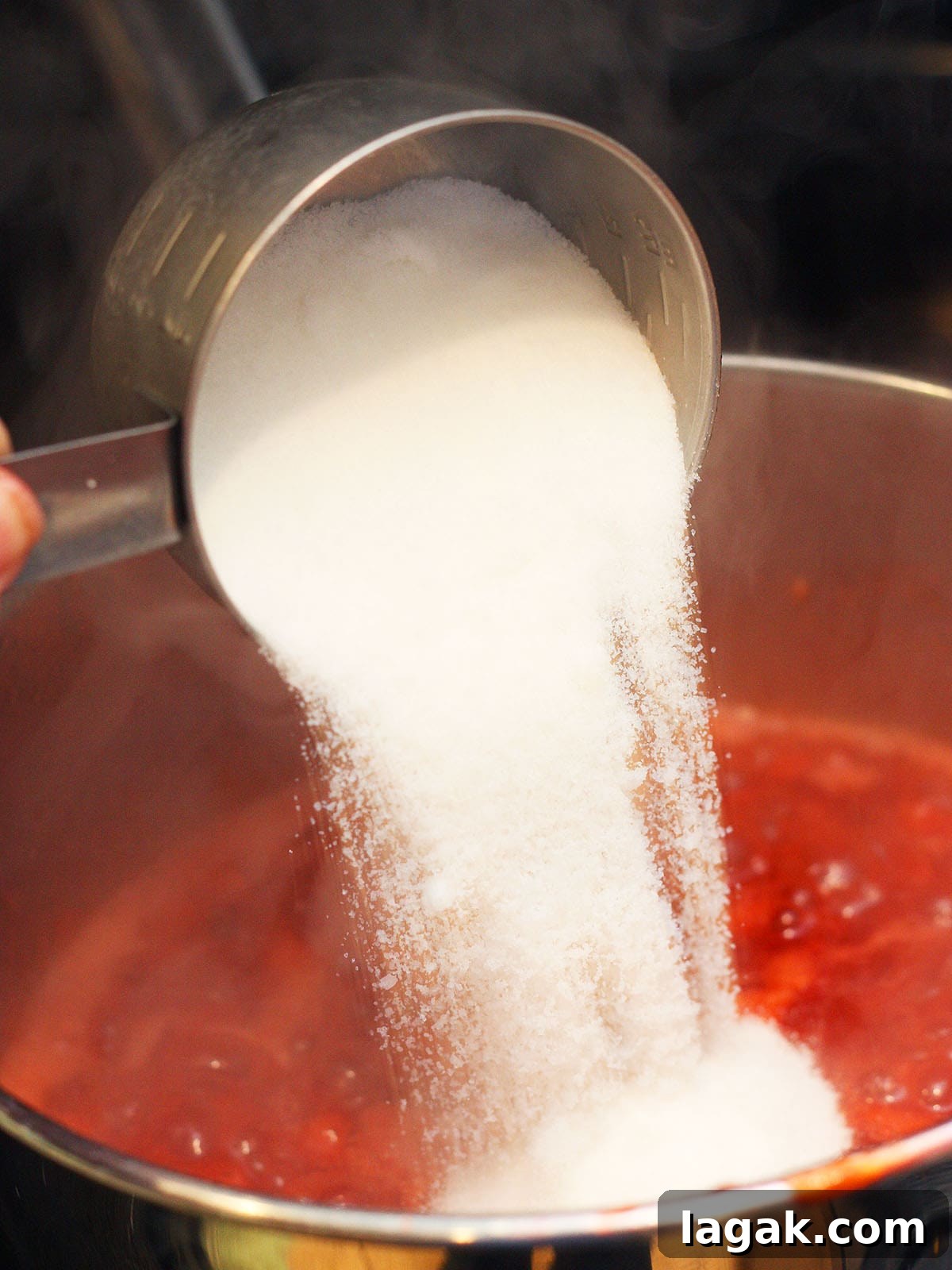 Strawberry Jam Recipe with Pectin: Smooth, Spreadable Preserves 8 Metal measuring cup pouring white sugar into a pot of strawberry preserves.
