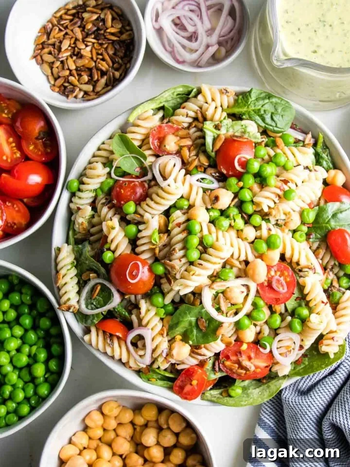 A white bowl filled with pasta salad and small bowls of the ingredients surrounding it: chick peas, green peas, halved cherry tomatoes, sunflower seeds, shallots and tahini parsley dressing.