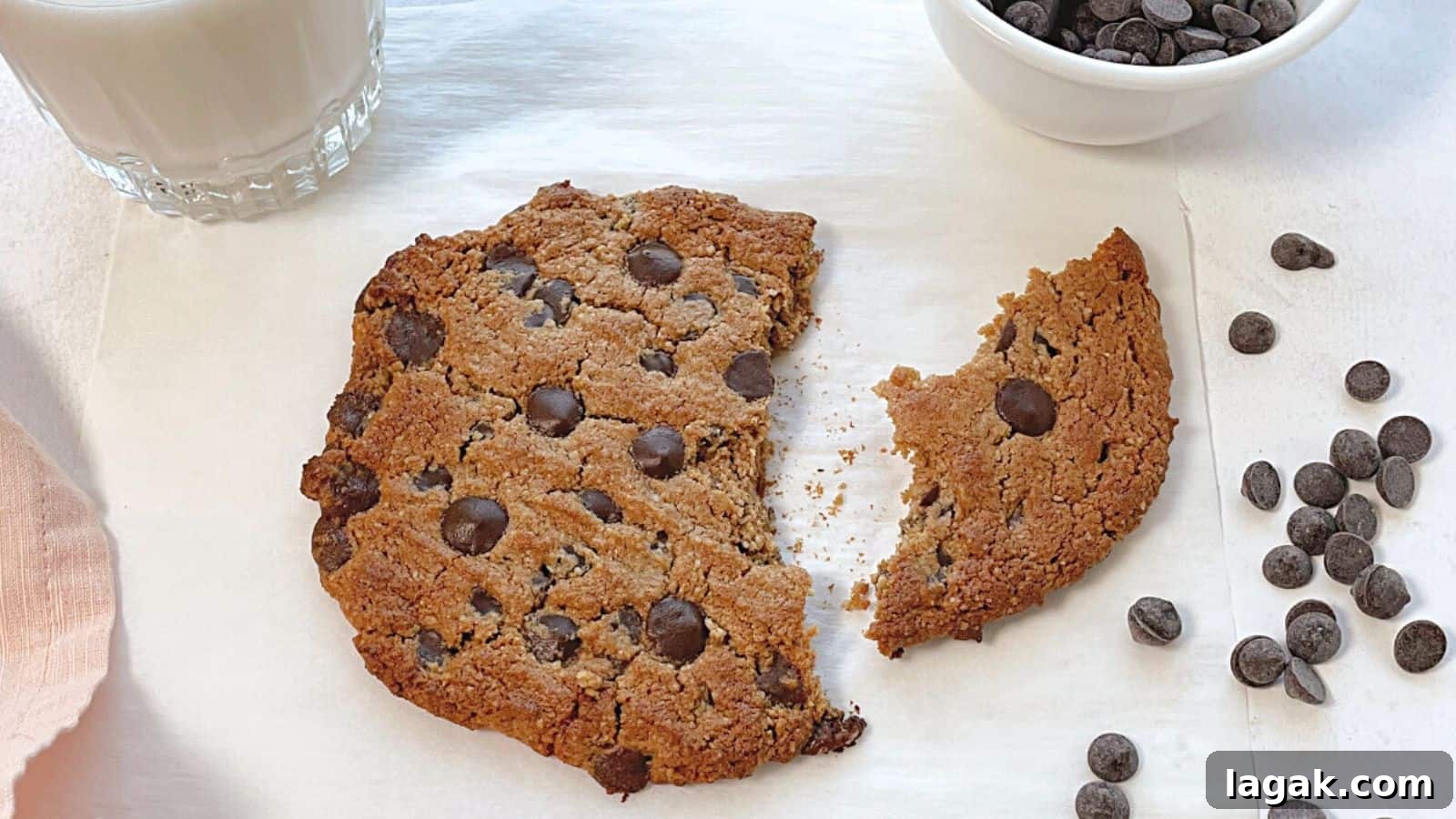 29 Party-Worthy Treats That Won’t Leave Leftovers 11 A large chocolate chip cookie with a piece broken off, next to a glass of milk and a bowl of chocolate chips on a white surface.