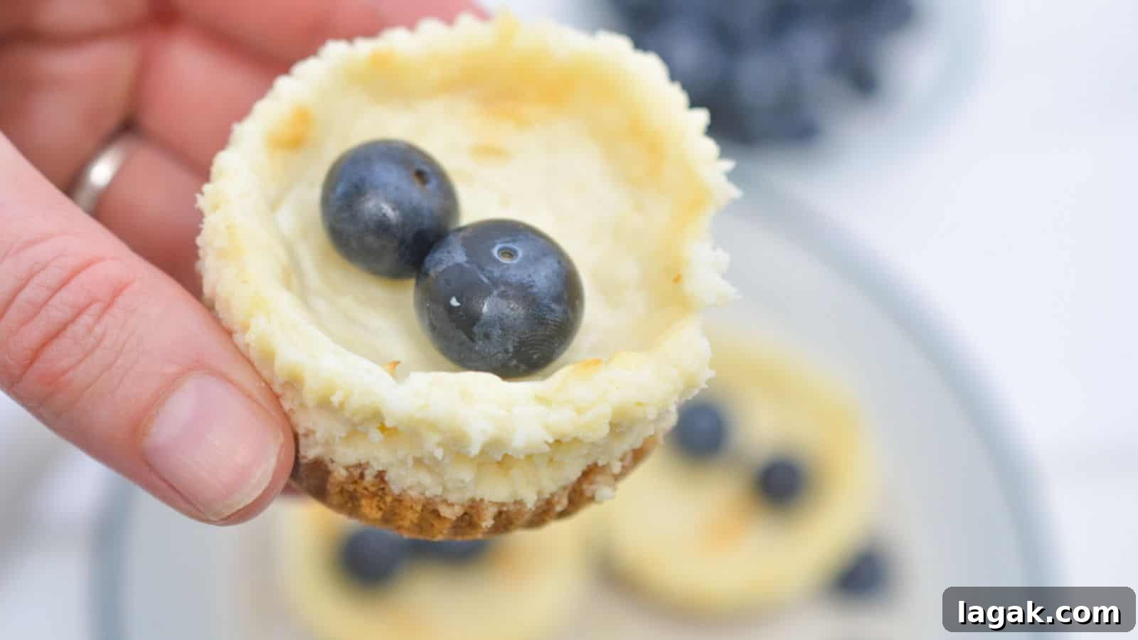 29 Party-Worthy Treats That Won’t Leave Leftovers 3 A hand holds a mini cheesecake topped with two blueberries, with more cheesecakes and berries on a plate in the background.