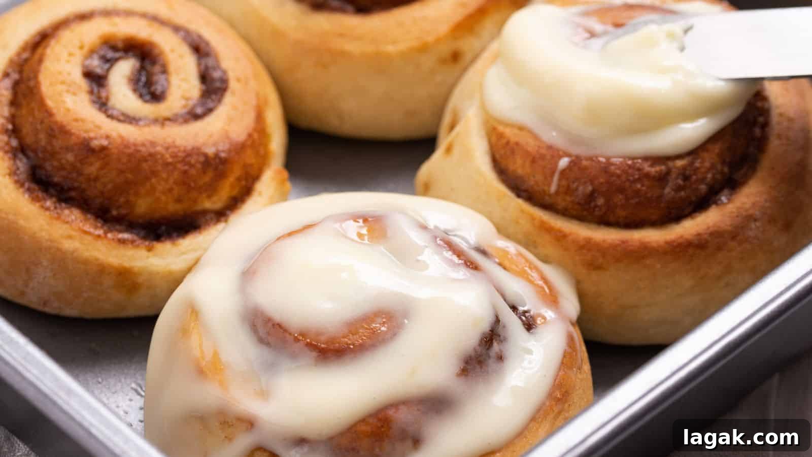29 Party-Worthy Treats That Won’t Leave Leftovers 22 A person spreads white frosting on a freshly baked cinnamon roll in a tray with others nearby.