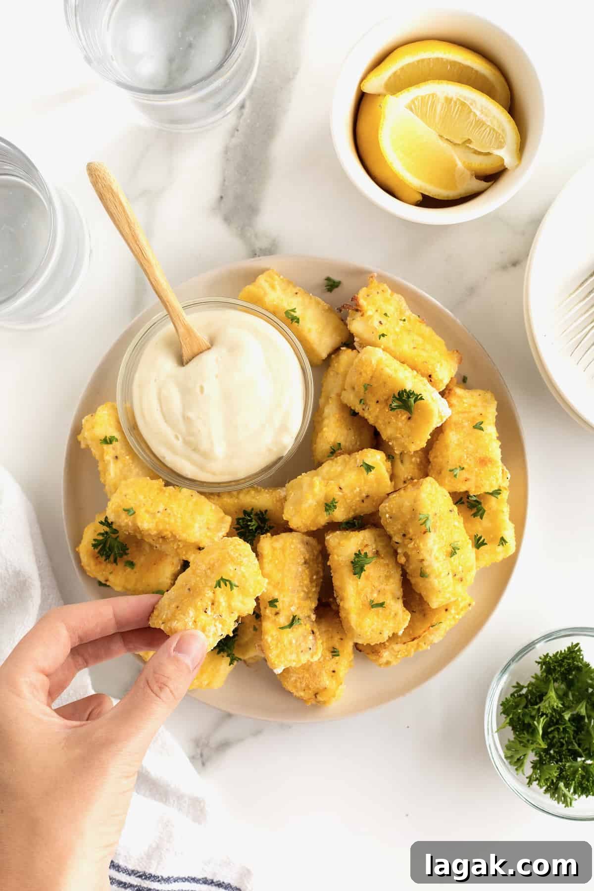 Crispy Oven-Baked Fish Sticks with Lemon Garlic Dip 12 A large white serving plate with a pile of golden battered fish sticks surrounding a small glass dish of tartar sauce. A hand is holding a fish stick over the platter.