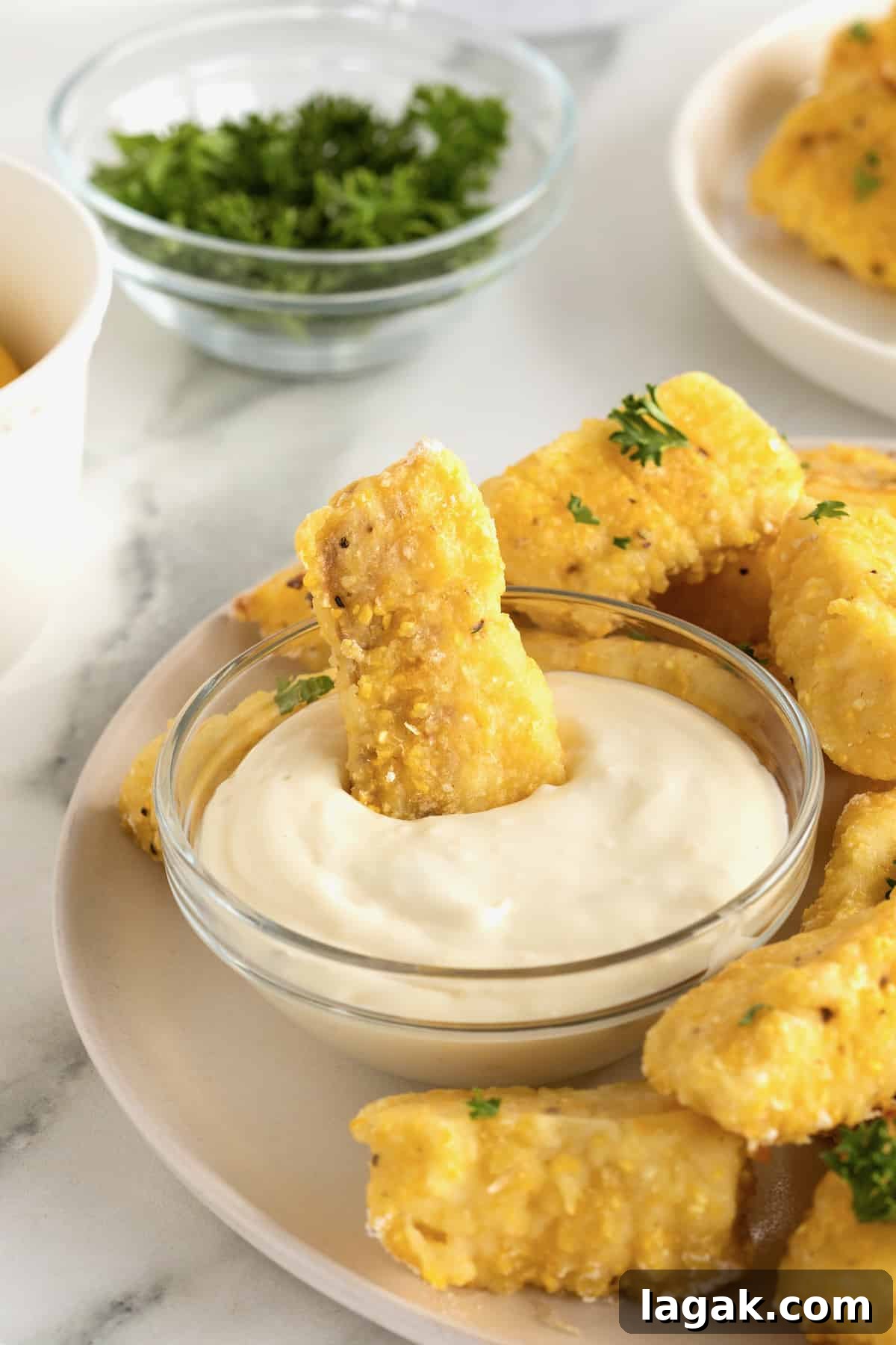 Crispy Oven-Baked Fish Sticks with Lemon Garlic Dip 5 A large white serving plate with a pile of golden battered fish sticks surrounding a small glass dish of tartar sauce. One fish stick is dipped into the tartar sauce.