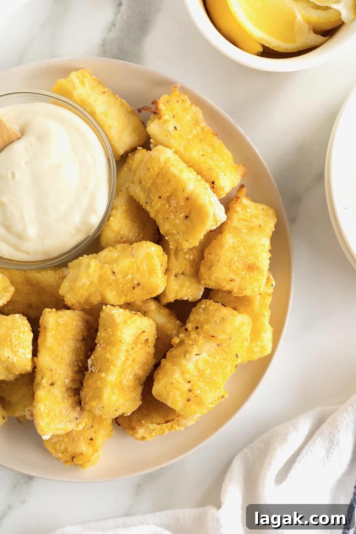 Crispy Oven-Baked Fish Sticks with Lemon Garlic Dip 6 A large white serving plate with a pile of golden battered fish sticks surrounding a small glass dish of tartar sauce.