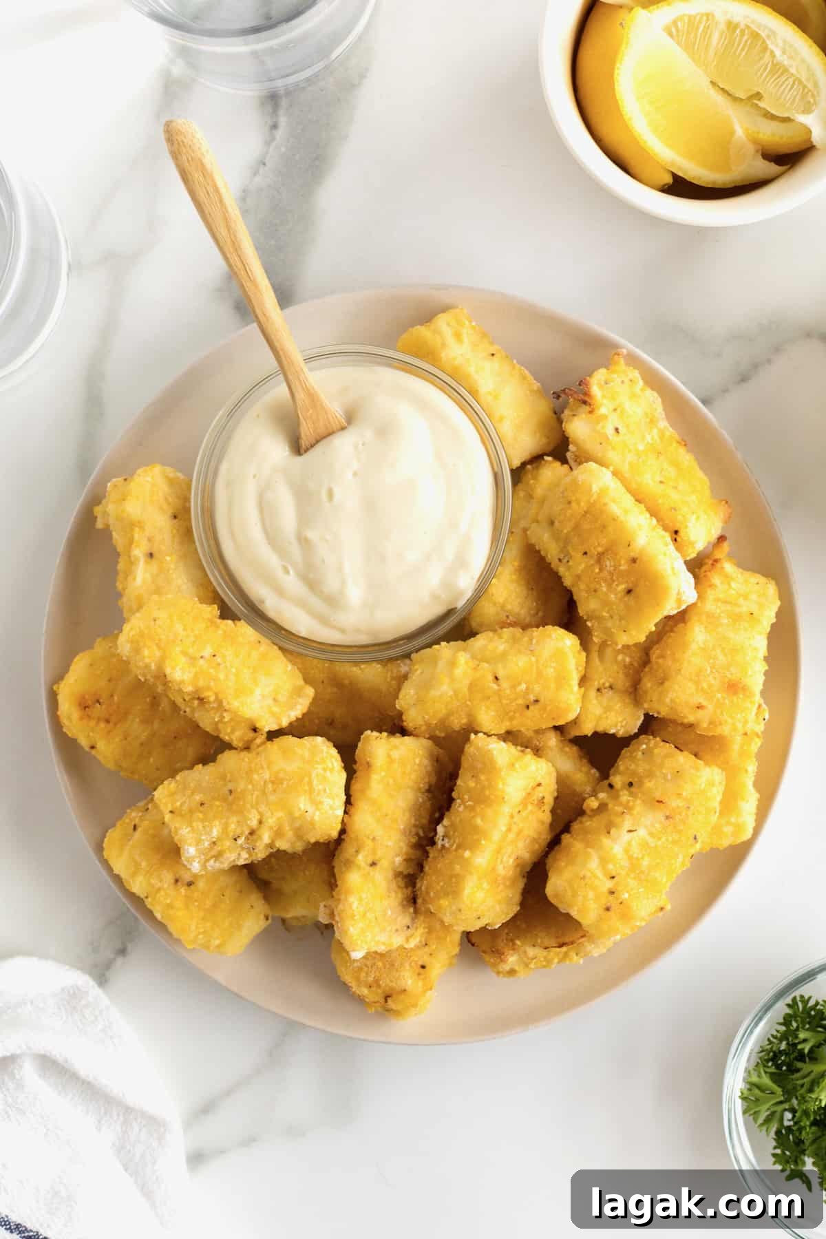 Crispy Oven-Baked Fish Sticks with Lemon Garlic Dip 7 A large white serving plate with a pile of golden battered fish sticks surrounding a small glass dish of tartar sauce. There's a small wooden spoon sticking out of the tartar sauce.