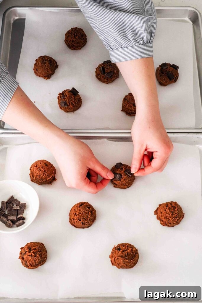 Ultimate Double Chocolate Chip Cookie Recipe for Rich, Gooey Cookies 11 Two hands push more chocolate chips onto cookie dough balls on baking sheets.