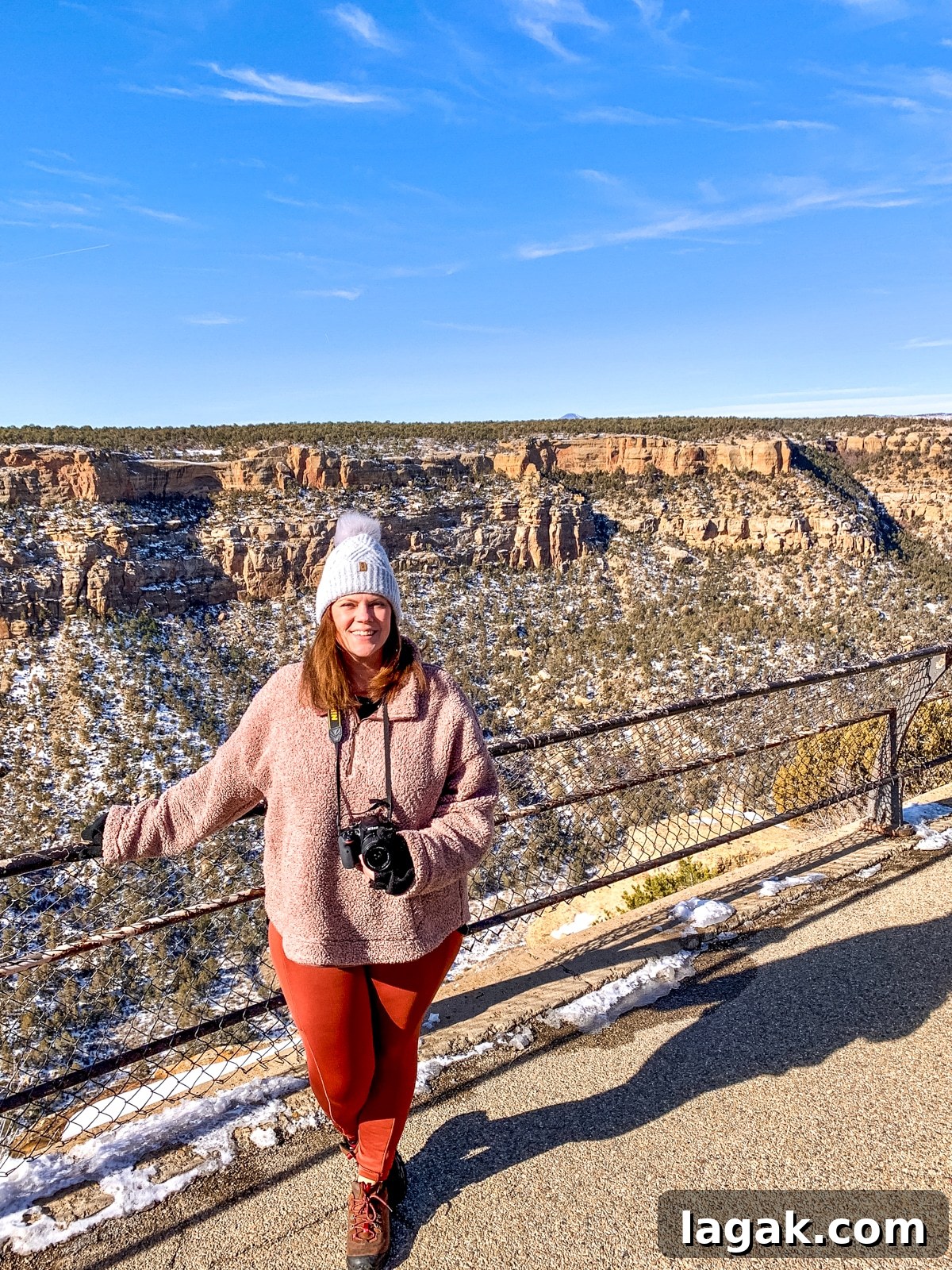Avoid Saying This When Planning Your Next Trip: Travel Tips 2 A person in a pink jacket and white beanie standing on a viewpoint overlooking a rocky, snowy canyon, holding a camera. The sky is clear and blue.