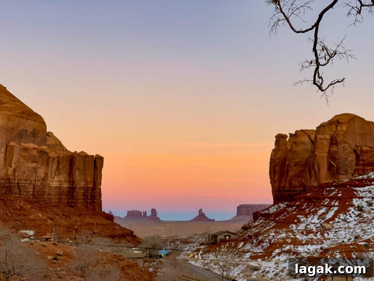 Avoid Saying This When Planning Your Next Trip: Travel Tips 3 A scenic view of Monument Valley at sunset, featuring large rock formations, a gradient colorful sky, and some snow on the ground. A few small buildings and a road are visible in the foreground.