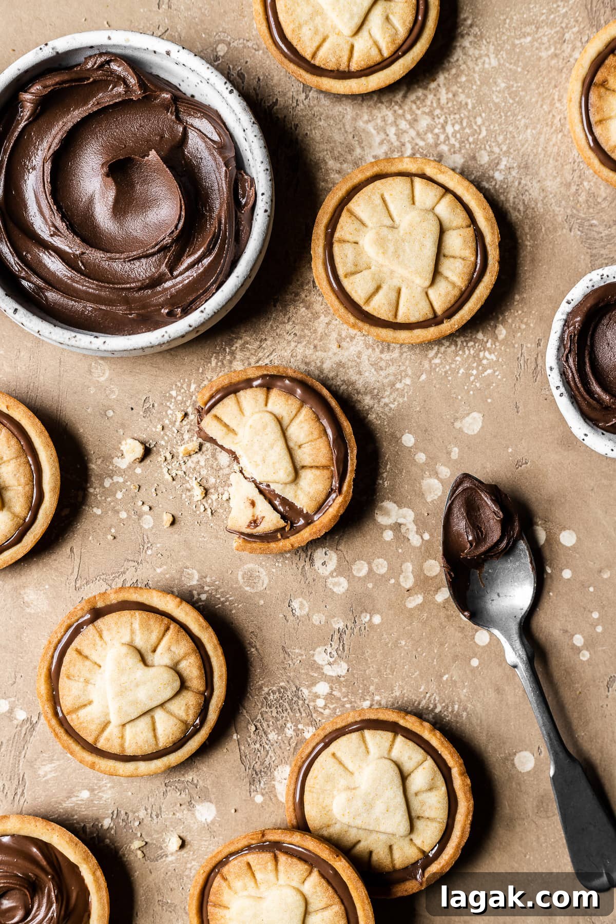 Homemade Nutella Biscuit Recipe: Crispy Chocolate Hazelnut Cookies 11 Nutella stuffed cookies on a warm tan speckled stone surface. A small white ceramic bowl is filled with a swirl of Nutella. An antique silver spoon with a scoop of Nutella rests on the surface at bottom right.