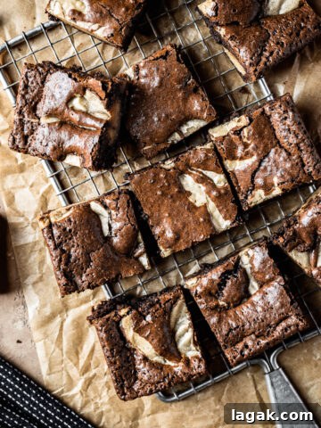 Brownie blondie squares casually arranged on a cooling rack with brown parchment underneath. A bowl of chocolate chips, a small plate with a brownie on top, and a dark blue napkin peek into the frame. The background surface is textured tan stone.