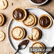 Homemade Nutella Biscuit Recipe: Crispy Chocolate Hazelnut Cookies 16 Nutella filled biscuits on a white speckled ceramic plate. A small bowl of Nutella rests nearby. A spoon of Nutella rests on the plate. The plate sits on a warm tan stone surface. More cookies and crumbs surround the plate.