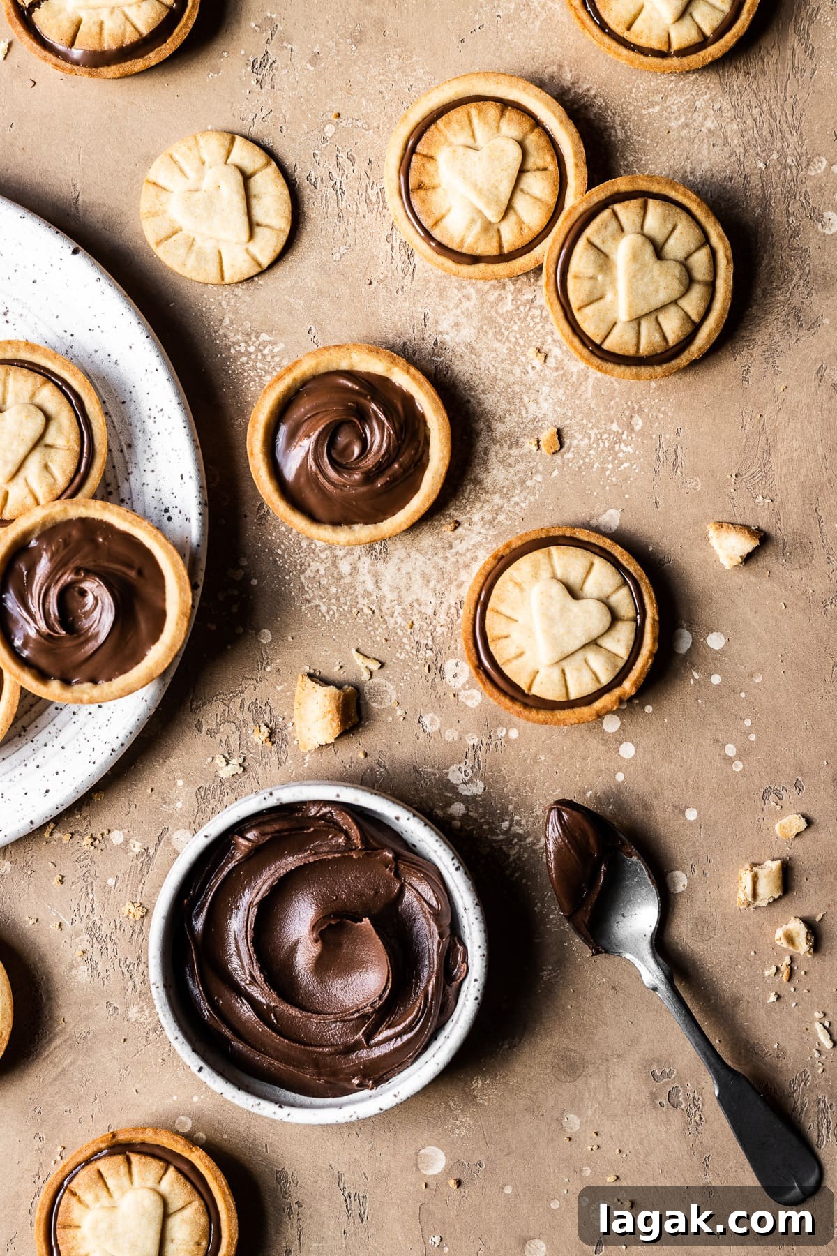 Homemade Nutella Biscuit Recipe: Crispy Chocolate Hazelnut Cookies 9 Chocolate hazelnut filled cookies on a warm tan stone surface. A small white ceramic bowl of Nutella rests at bottom with a silver spoon nearby. A white ceramic plate full of cookies peeks into the photo at left.