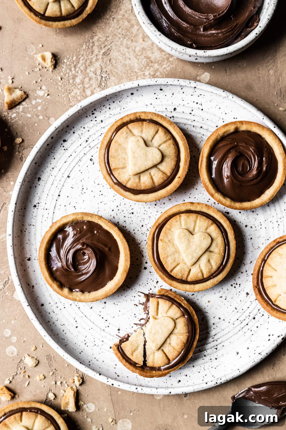 Homemade Nutella Biscuit Recipe: Crispy Chocolate Hazelnut Cookies 10 Golden brown honey Nutella biscuits on a white speckled ceramic plate. A small bowl of Nutella rests nearby. The plate sits on a warm tan stone surface. More cookies and crumbs surround the plate.