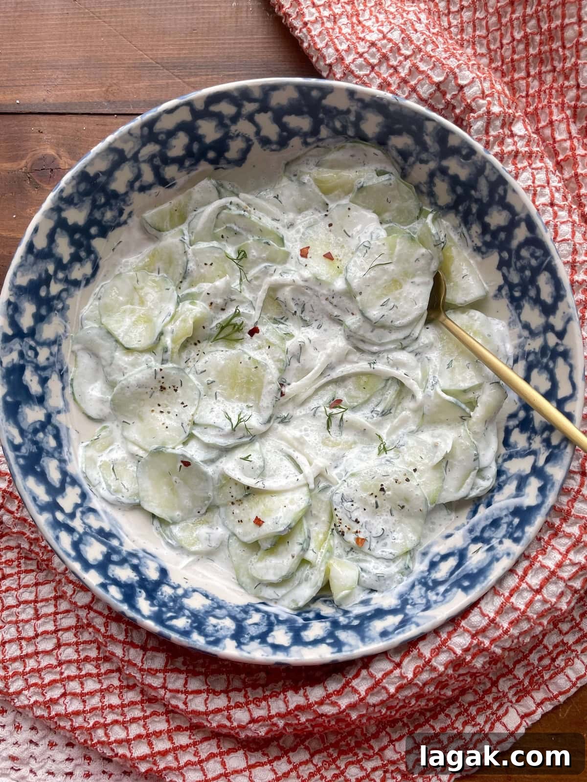 Creamy German Gurkensalat Recipe: Classic Cucumber Salad with Dill 2 A blue bowl of creamy cucumber salad with sliced onions, fresh dill, and red pepper flakes and a gold spoon.