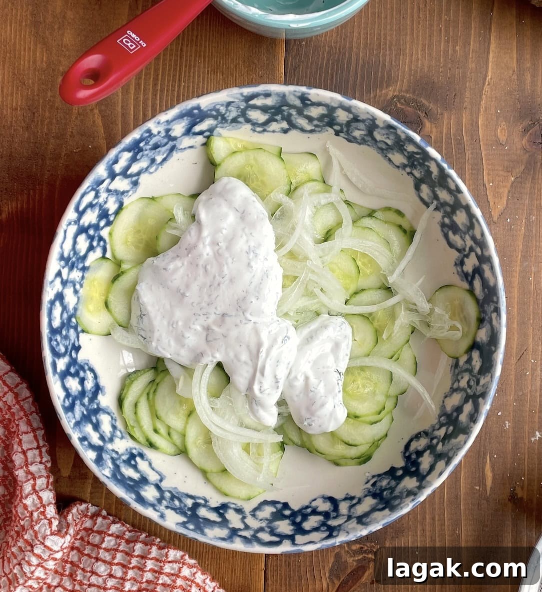 Creamy German Gurkensalat Recipe: Classic Cucumber Salad with Dill 8 A bowl of cucumbers and onions with creamy sauce on top before it is mixed together.