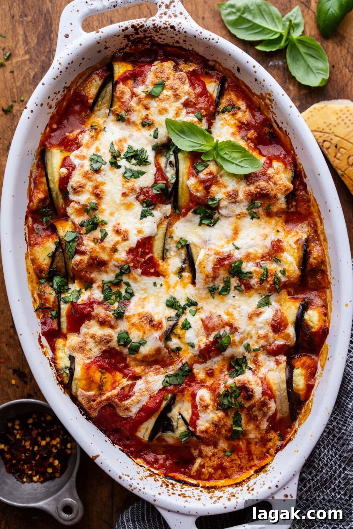 Classic Eggplant Rollatini Recipe — Simple Italian Comfort Food 2 A white dish with eggplant rollatini in it, topped with fresh herbs. The dish is sitting on a wooden background with a cheese rind, napkin, and a small bowl of red pepper flakes arranged around it.