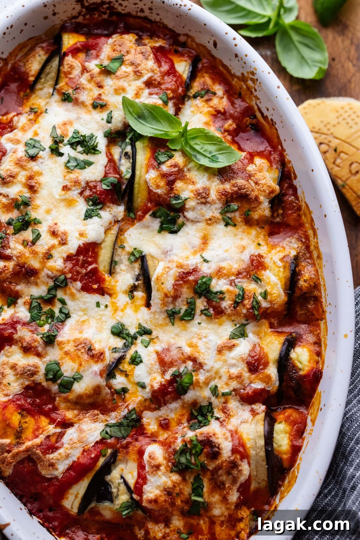 Classic Eggplant Rollatini Recipe — Simple Italian Comfort Food 6 Eggplant rollatini with fresh herbs sprinkled over top in a white dish. Next to the dish is a parmesan cheese rind and fresh herbs.