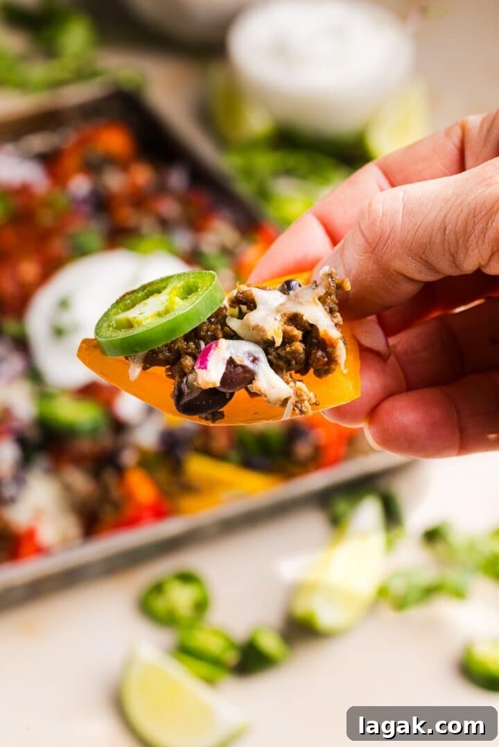 Bell Pepper Nachos with Zesty Cheese and Fresh Toppings 10 Woman's hand holding a bell pepper nacho up to the camera, with a full sheet tray of loaded nachos behind her.